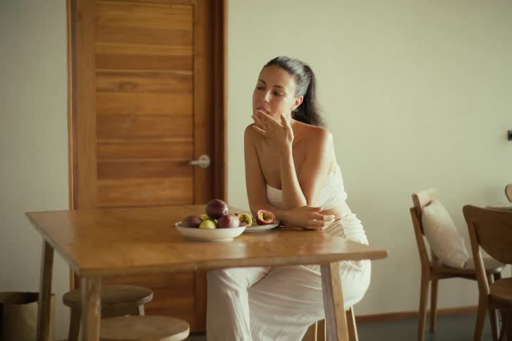 A happy woman enjoys a bite of fresh fruit while sitting at a wooden table, then smiles and laughs joyfully at the camera