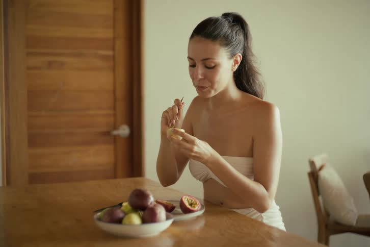 A beautiful young woman in a white top enjoys eating a fresh passion fruit with a spoon in slow motion