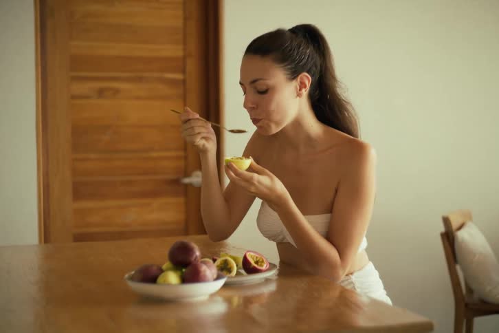 A beautiful young woman in slow motion eats a fresh passion fruit with a spoon at her breakfast table