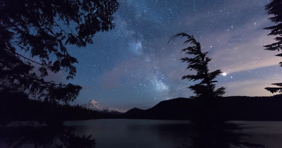 A breathtaking timelapse shows the Milky Way and meteors streaking across the night sky over a tranquil mountain lake before dawn breaks