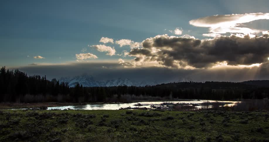A stunning timelapse shows sun rays piercing through moving clouds over a tranquil river, forest, and distant snow-capped mountains