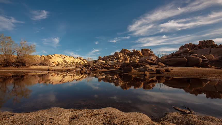 A tranquil timelapse of wispy clouds reflecting in a still desert pond surrounded by sandy rock formations