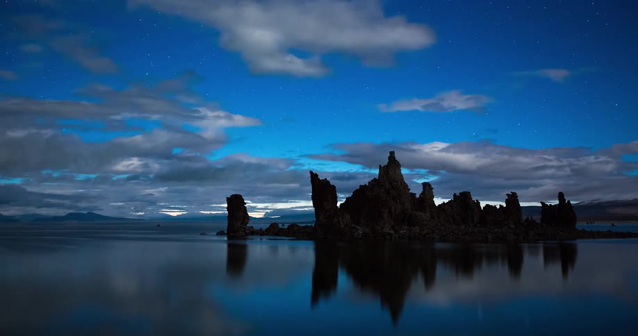 A tranquil time-lapse shows clouds moving across a starry night sky over the tufa towers of Mono Lake, perfectly reflected in the calm water