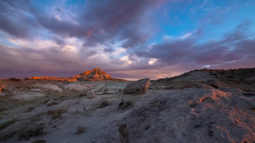 A majestic time-lapse shows dramatic purple and pink clouds moving across the sky at sunset over a rocky desert badlands