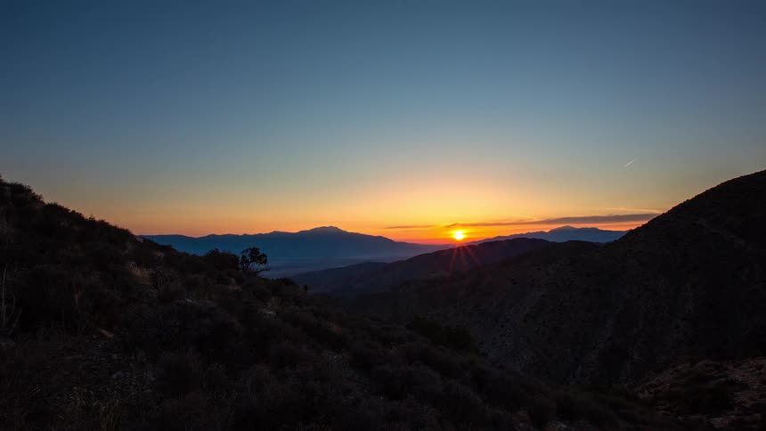 A scenic timelapse captures the sun setting behind a vast mountain range, transitioning the landscape from day to night