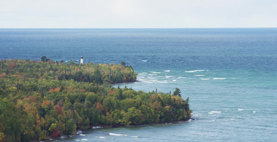 A distant lighthouse peeks through a vibrant autumn forest along the rugged coastline of a vast blue lake