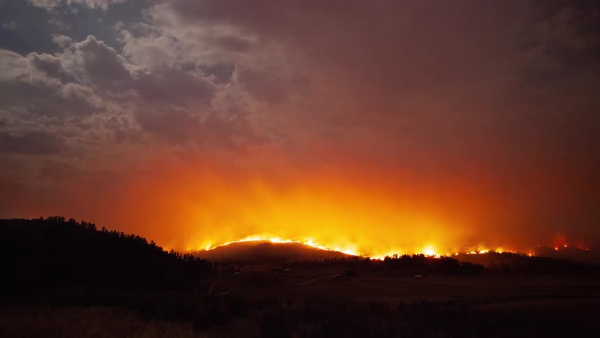 A dramatic time-lapse of a massive wildfire raging across a hillside at night, casting an apocalyptic orange glow into the cloudy sky