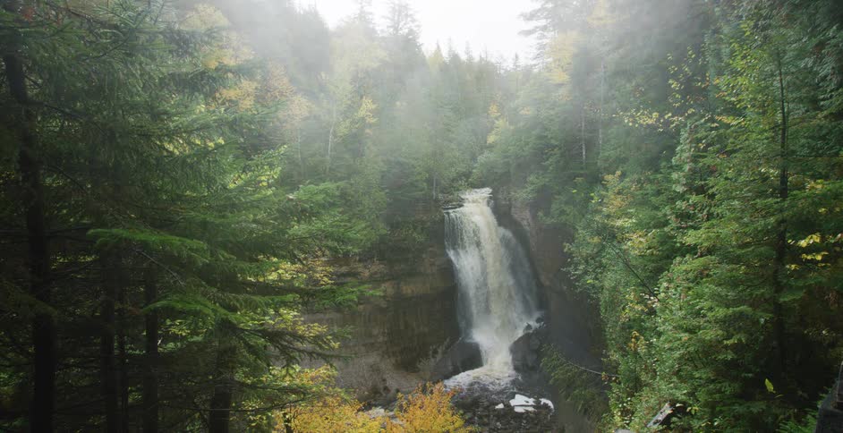 A serene waterfall cascades down a rocky cliff in a dense, foggy forest during autumn