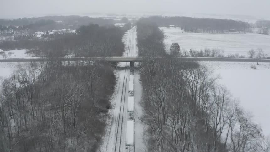 An aerial view shows a freight train traveling through a snowy, tree-lined rural landscape and under a highway bridge during a winter storm