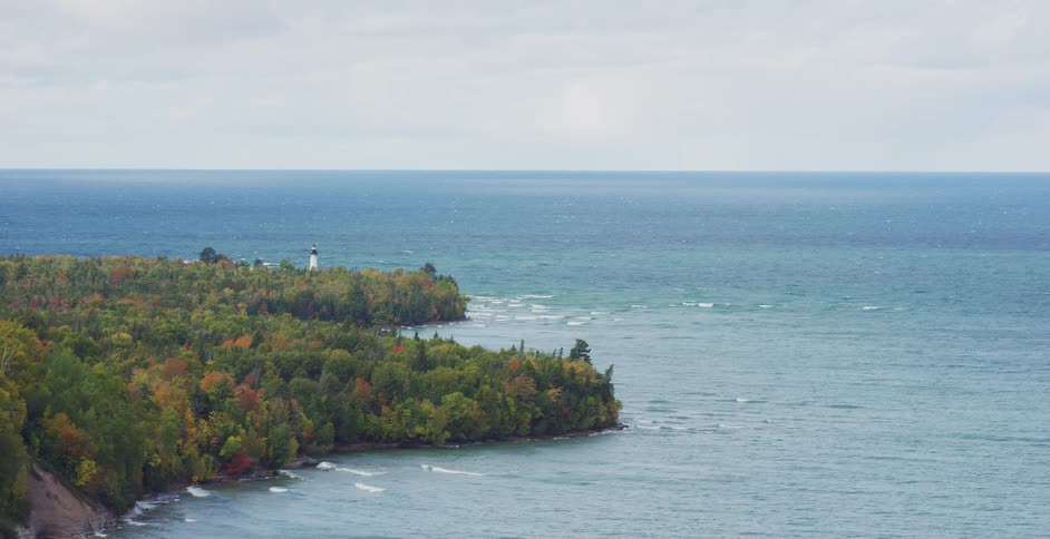 A scenic wide shot of a lighthouse on a forested peninsula with autumn colors, overlooking a vast blue lake on an overcast day