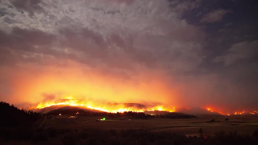 A dramatic night time-lapse shows a massive wildfire raging across a mountain range, illuminating the smoky sky with an ominous orange glow