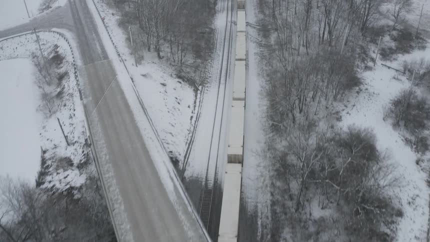 High angle aerial shot of a freight train traveling through a stark, snow-covered rural landscape during a winter storm