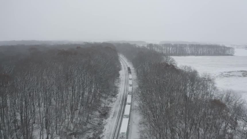 Slow aerial shot follows a freight train as it moves through a vast, snowy woodland landscape during a winter storm