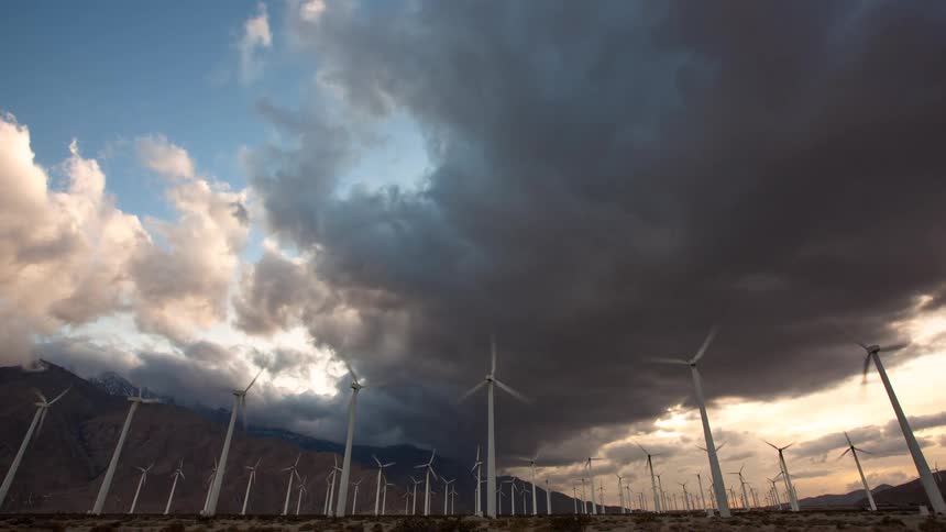 Time-lapse of a massive wind farm with spinning turbines against a mountain range as dark storm clouds gather at dusk