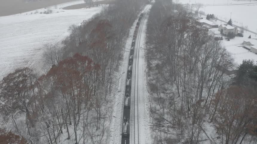 An aerial view slowly pulls back from a long freight train traveling through a snow-covered forest during a winter day