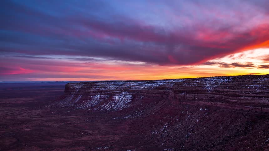 A breathtaking time-lapse captures a vibrant sunset painting the sky with brilliant colors over a snow-dusted desert mesa