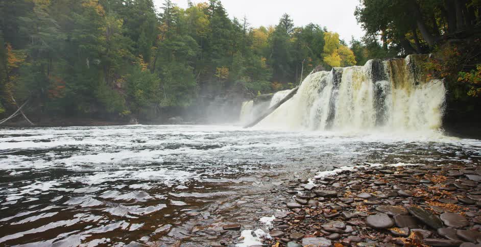 A wide, powerful waterfall thunders over a rocky cliff into a river, surrounded by a misty forest showing early signs of autumn