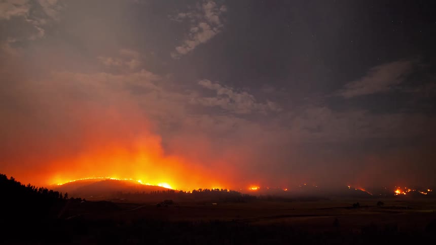 A massive wildfire burns intensely on a hillside at night, with smoke billowing into the starry, cloud-filled sky in a dramatic time-lapse
