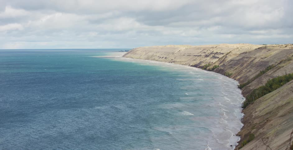 Gentle waves from a vast blue lake crash against the base of towering sand dunes on a cloudy day