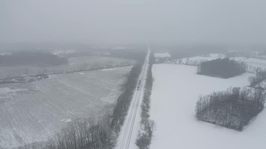 An aerial view shows a freight train traveling through a rural, snow-covered landscape during a heavy snowstorm