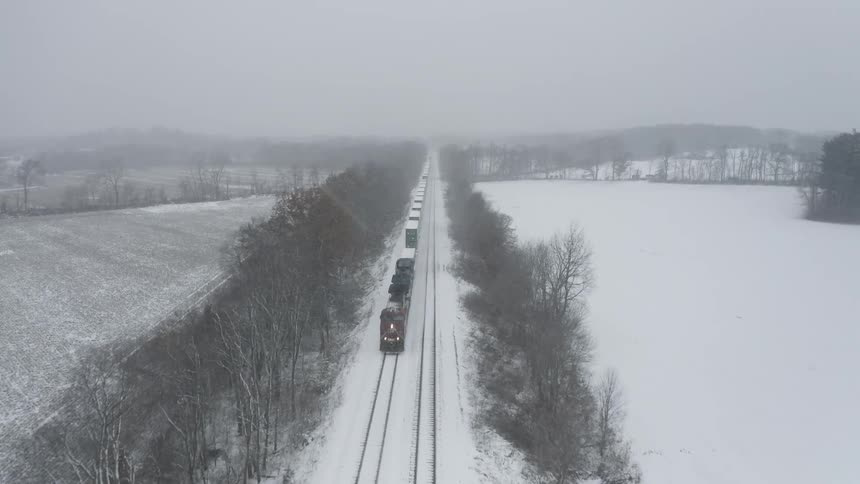 From a high aerial perspective, a freight train approaches on a long, straight track that cuts through a vast, snow-covered rural landscape during a winter storm