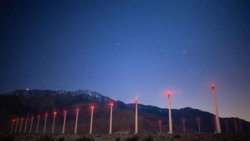 A stunning night time-lapse shows wind turbines spinning under a star-filled sky with snow-capped mountains in the background