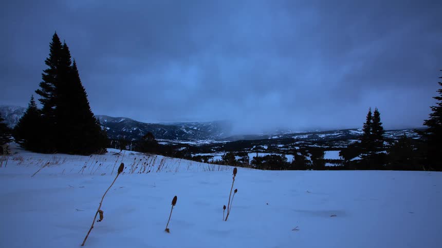 A time-lapse shows dark, moody clouds moving across a snow-covered mountain valley at twilight