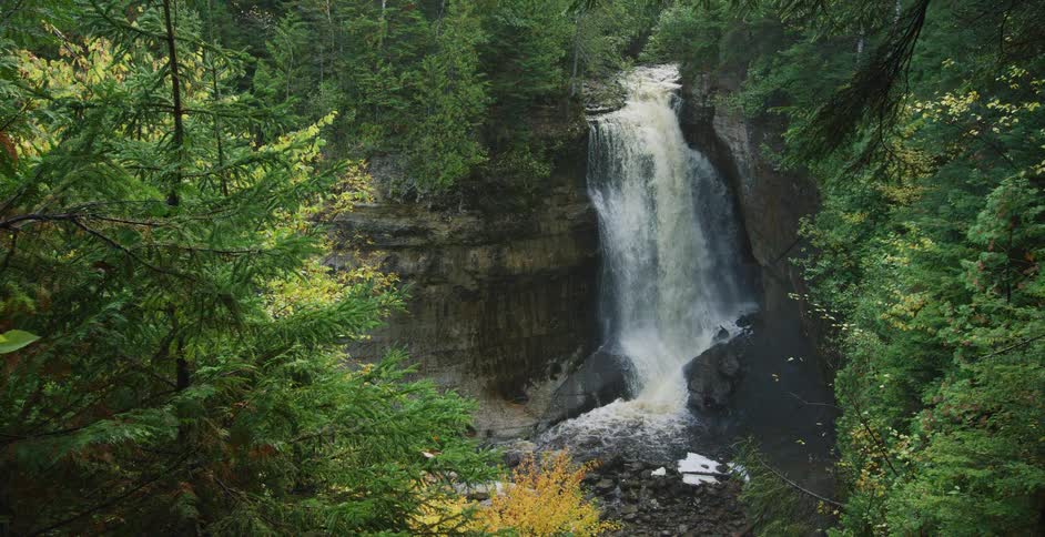 A powerful waterfall cascades down a rocky cliff in a lush green forest with hints of yellow autumn leaves