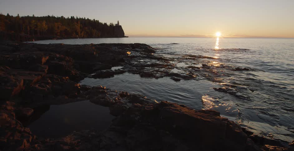 A serene sunrise casts a golden glow over a calm lake, illuminating the rocky shoreline and a distant lighthouse
