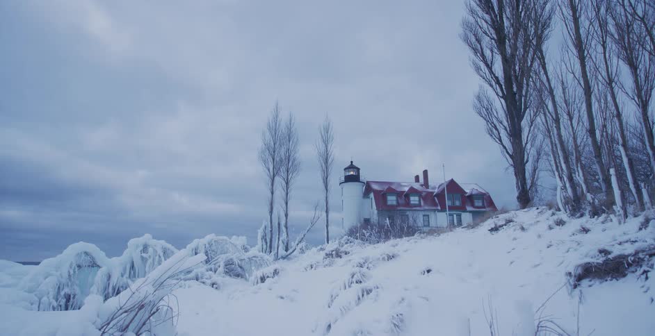 A historic lighthouse sits atop a snow-covered hill on a cold, overcast winter day by a frozen lake