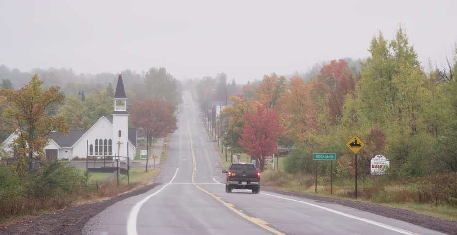 A pickup truck drives down a foggy country road past a quaint white church amidst vibrant autumn foliage
