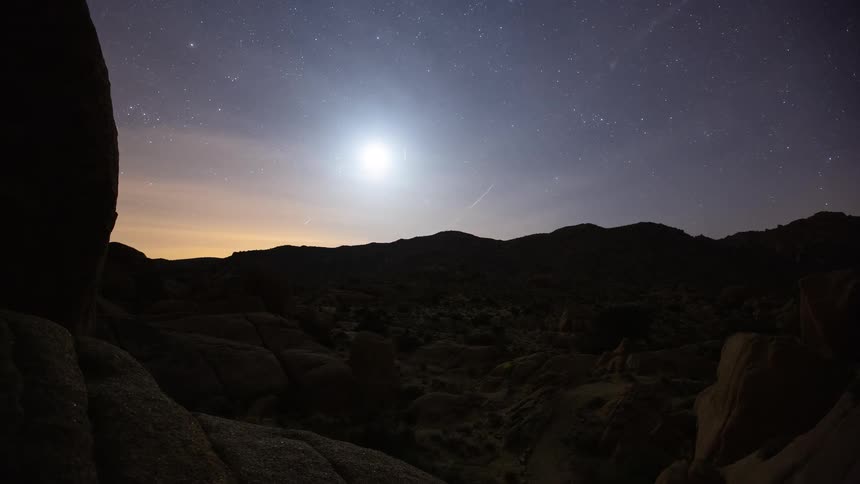 Time-lapse of the moon setting behind silhouetted mountains in the rocky desert as stars and meteors streak across the night sky