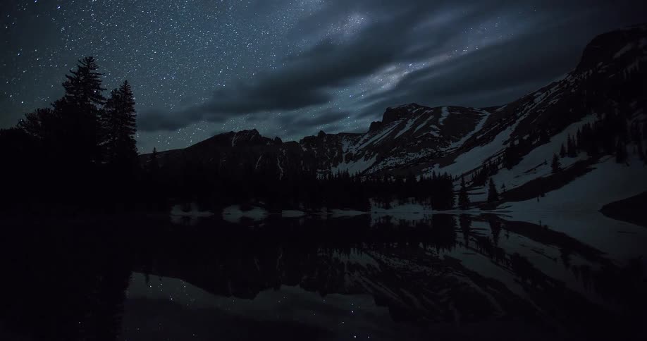 Breathtaking time-lapse from dusk to dawn captures the Milky Way and stars moving over a snowy mountain range reflected in a calm alpine lake