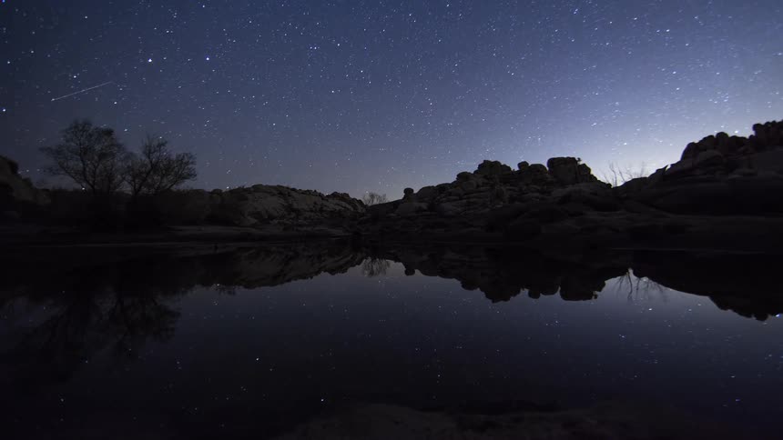 A breathtaking day-to-night timelapse captures the transition to a star-filled sky, perfectly reflected in a still desert lake surrounded by rocky formations