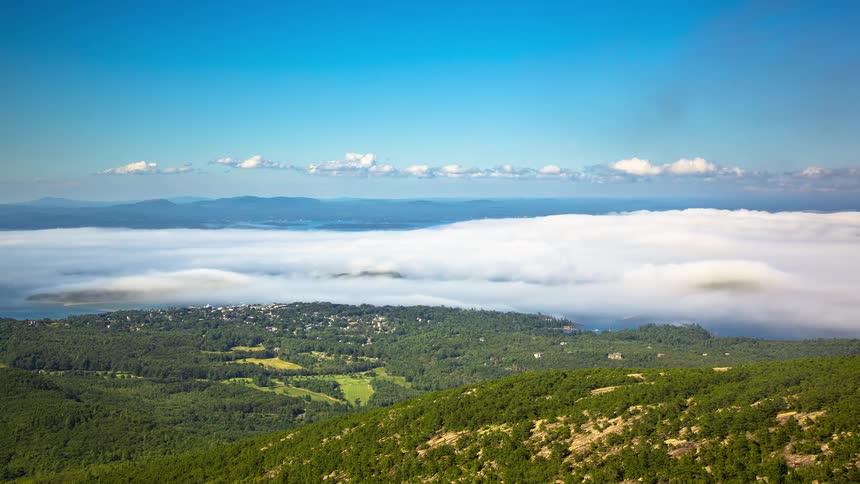 A timelapse of thick white fog rolling over a coastal town and islands as viewed from a mountaintop on a clear day