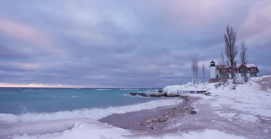 Slow-motion waves crash onto a snowy, ice-covered shoreline next to a lighthouse on a cold, cloudy winter day