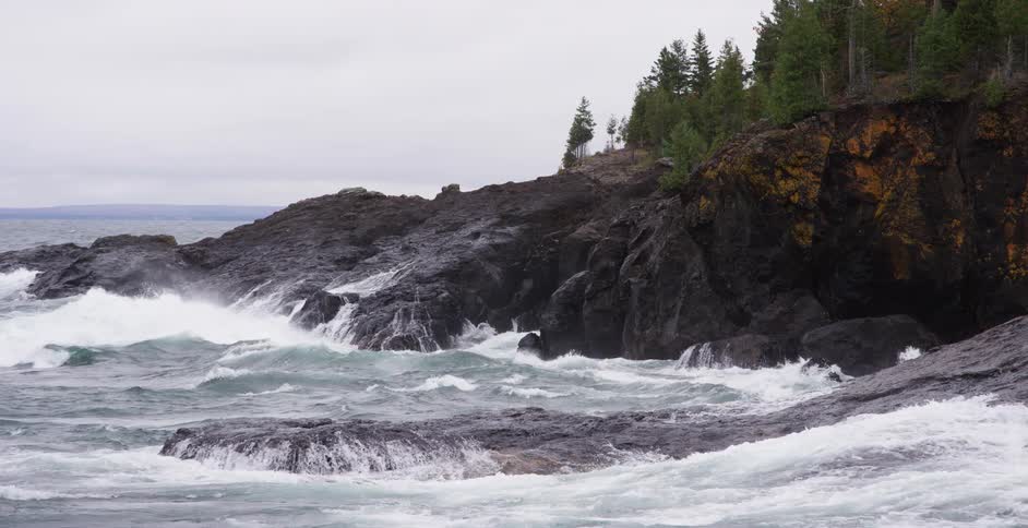 Powerful waves crash against a rocky, tree-lined coastline in dramatic slow motion on an overcast day