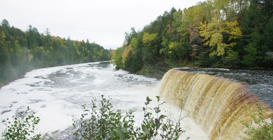 A wide, powerful waterfall tumbles into a churning river, surrounded by a dense forest displaying beautiful early autumn colors