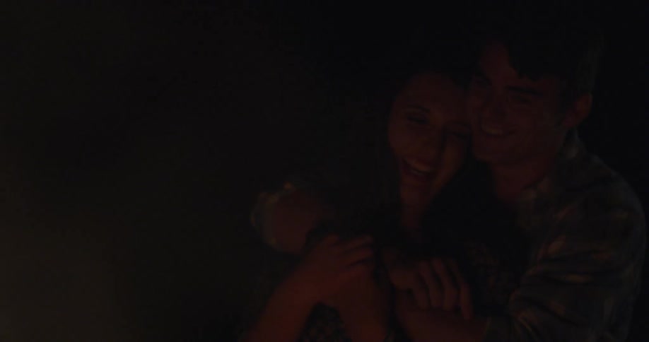 Man and woman sit in front of a fire smiling at night