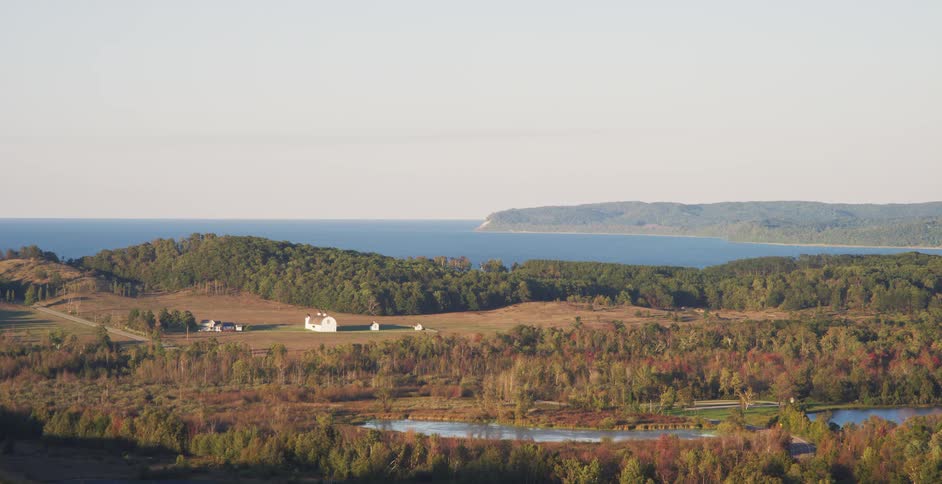 A serene, wide landscape of a rural farm and autumn-colored forests overlooking a large lake with distant sand dunes