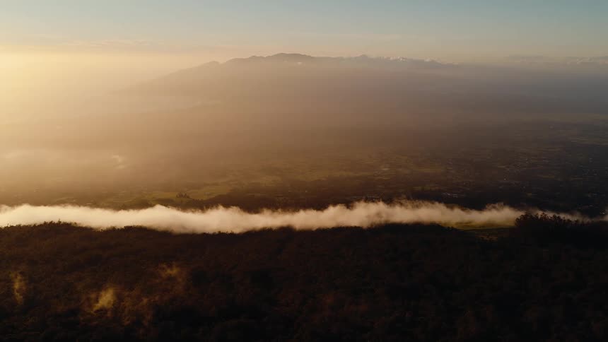 A stunning aerial view of a golden sunrise over a hazy mountain valley, with a long line of clouds separating the dark forest from the landscape beyond