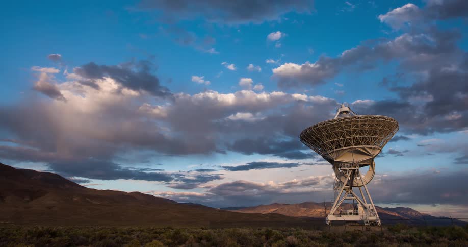 A dramatic sunset timelapse shows clouds moving over a large satellite dish in a remote desert valley