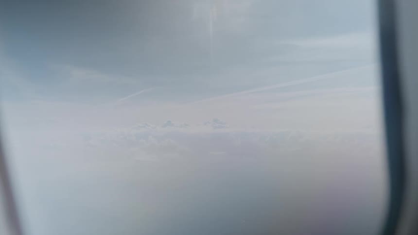 A serene aerial view of a hazy sky and clouds slowly zooms out from an airplane window