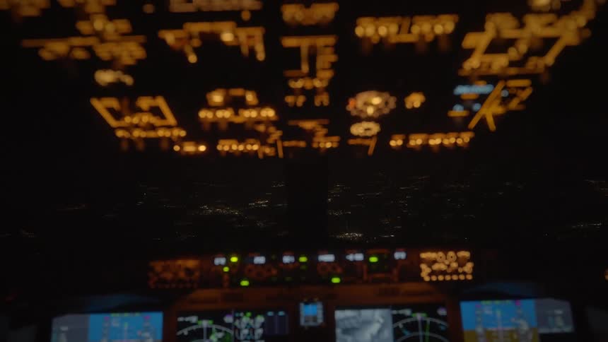 Blurred airplane cockpit ceiling lights glow over a sparkling cityscape seen through the windshield at night