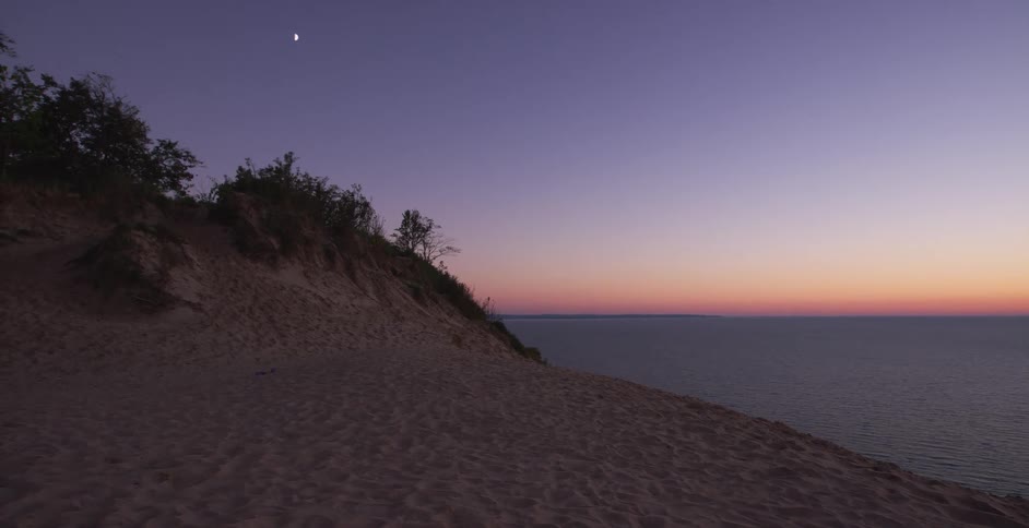 A crescent moon rises in the purple twilight sky over a calm ocean as seen from the top of a sand dune