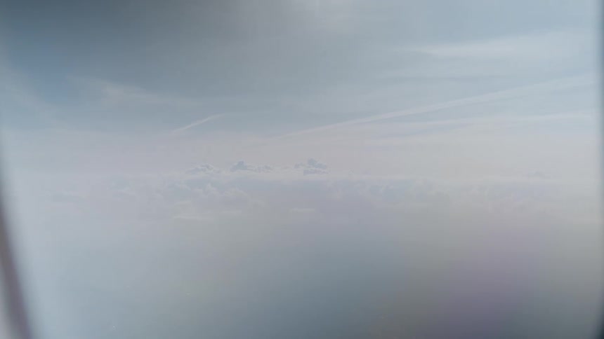 A smooth push-in shot from an airplane window reveals a vast, hazy sky above the clouds