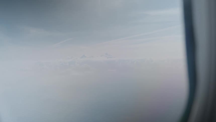 A serene view of the sky and clouds slowly zooms out from an airplane window during flight