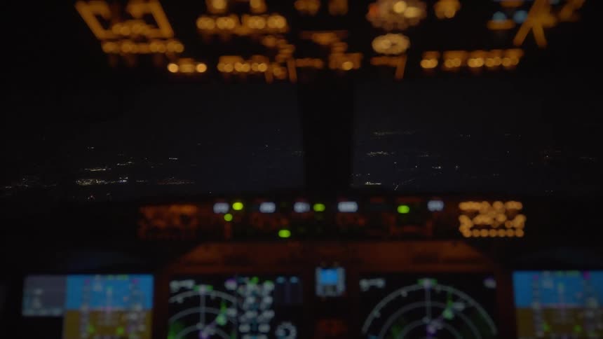 An out-of-focus shot from an airplane cockpit at night reveals the city lights twinkling below