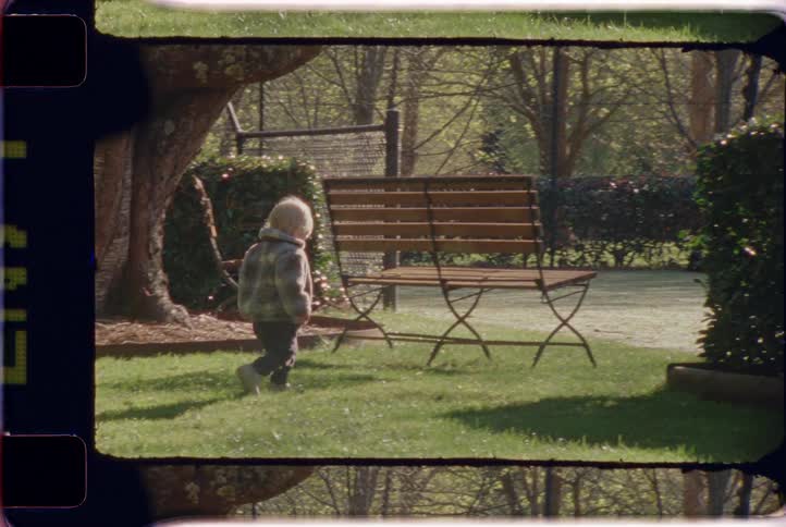 A small child in a plaid coat wanders through a sun-dappled park in this nostalgic, vintage-style footage
