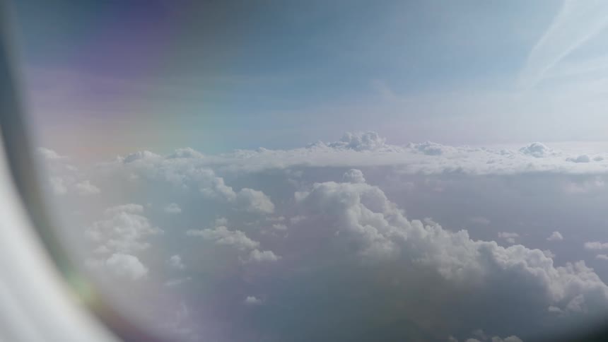 Dreamy aerial view of fluffy white clouds and blue sky as seen from an airplane window with a subtle rainbow lens flare